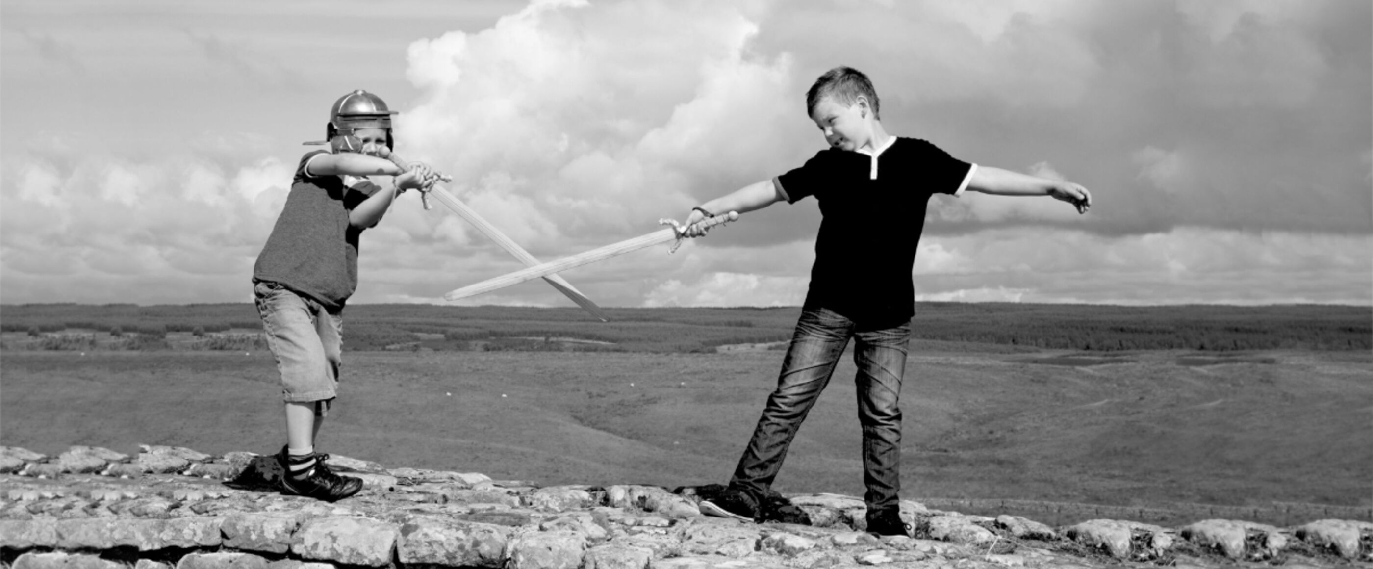 Zwei Kinder stehen mit Holzschwertern auf einer Mauer und spielen Krieg.