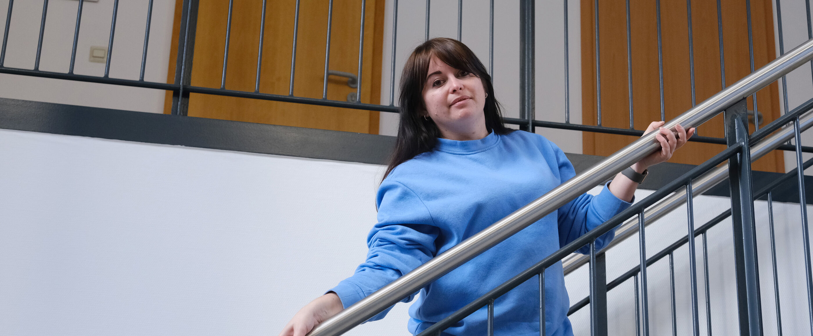 Eine als Frau lesbare Person mit dunklen Haaren und einem blauen Pullover steht an einem Treppengeländer.
