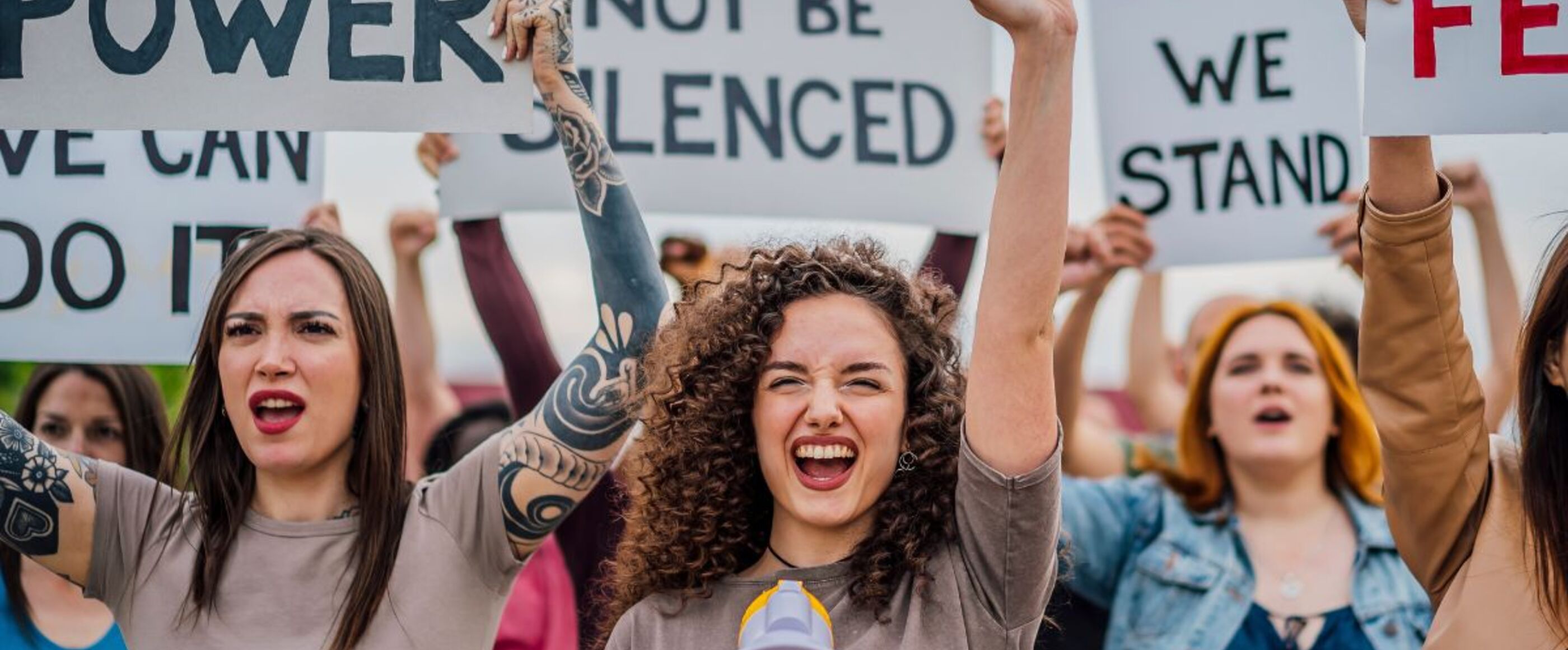 Junge, weiblich gelesene Personen demonstrieren mit Plakaten, auf denen steht: Girl power - Frauenpower, We will not be silenced - Wir werden nicht leise gestellt, United we stand - Wir stehen gemeinsam